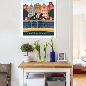 May include: A framed poster with a vintage travel illustration of Amsterdam, Netherlands. The poster features a couple riding a bicycle on a bridge with colorful buildings in the background. The text "AMSTERDAM" and "Netherlands" is at the top of the poster. The text "KATE & SCHMIDT" is at the bottom of the poster.