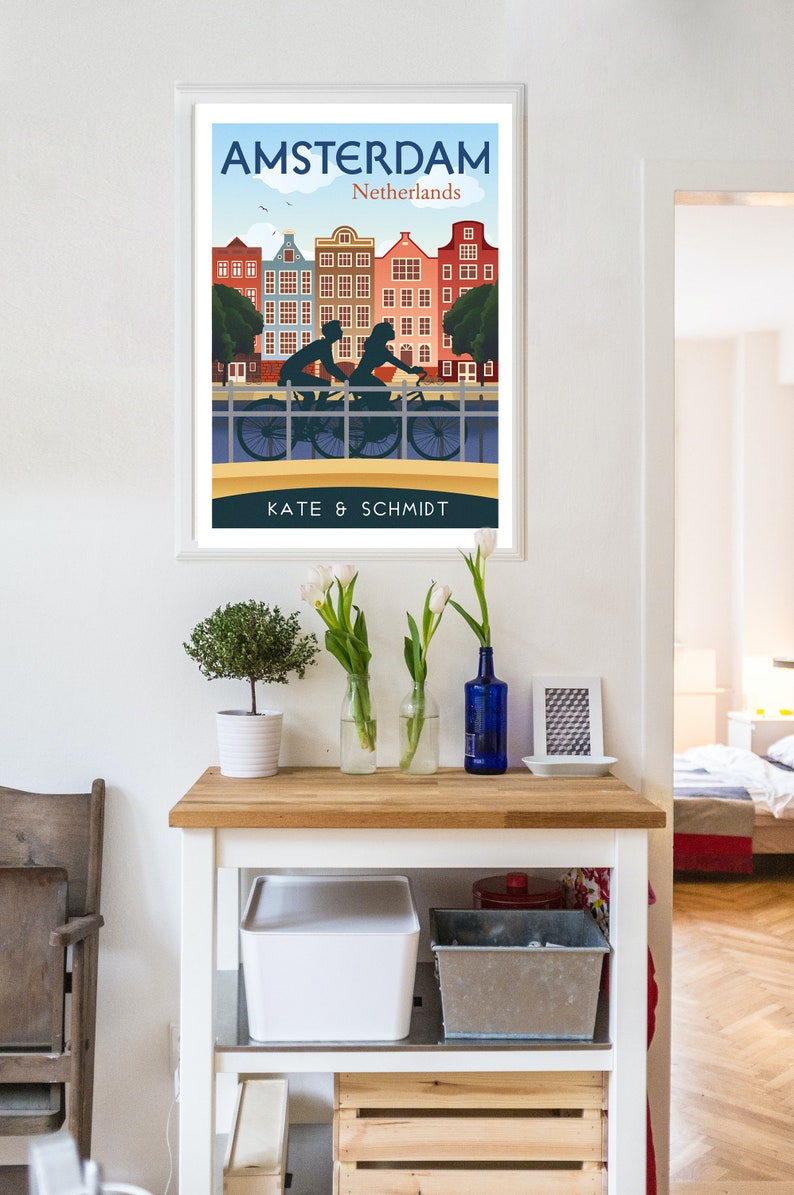 May include: A framed poster with a vintage travel illustration of Amsterdam, Netherlands. The poster features a couple riding a bicycle on a bridge with colorful buildings in the background. The text "AMSTERDAM" and "Netherlands" is at the top of the poster. The text "KATE & SCHMIDT" is at the bottom of the poster.