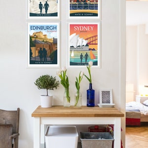 May include: Four framed travel posters with a couple silhouette in each. The posters are for Santorini, Greece; Amsterdam, Netherlands; Edinburgh, Scotland; and Sydney, Australia. The posters are hanging on a white wall with a wooden table below.