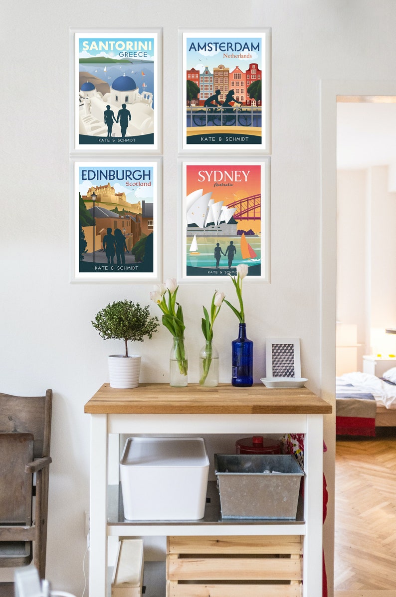 May include: Four framed travel posters with a couple silhouette in each. The posters are for Santorini, Greece; Amsterdam, Netherlands; Edinburgh, Scotland; and Sydney, Australia. The posters are hanging on a white wall with a wooden table below.