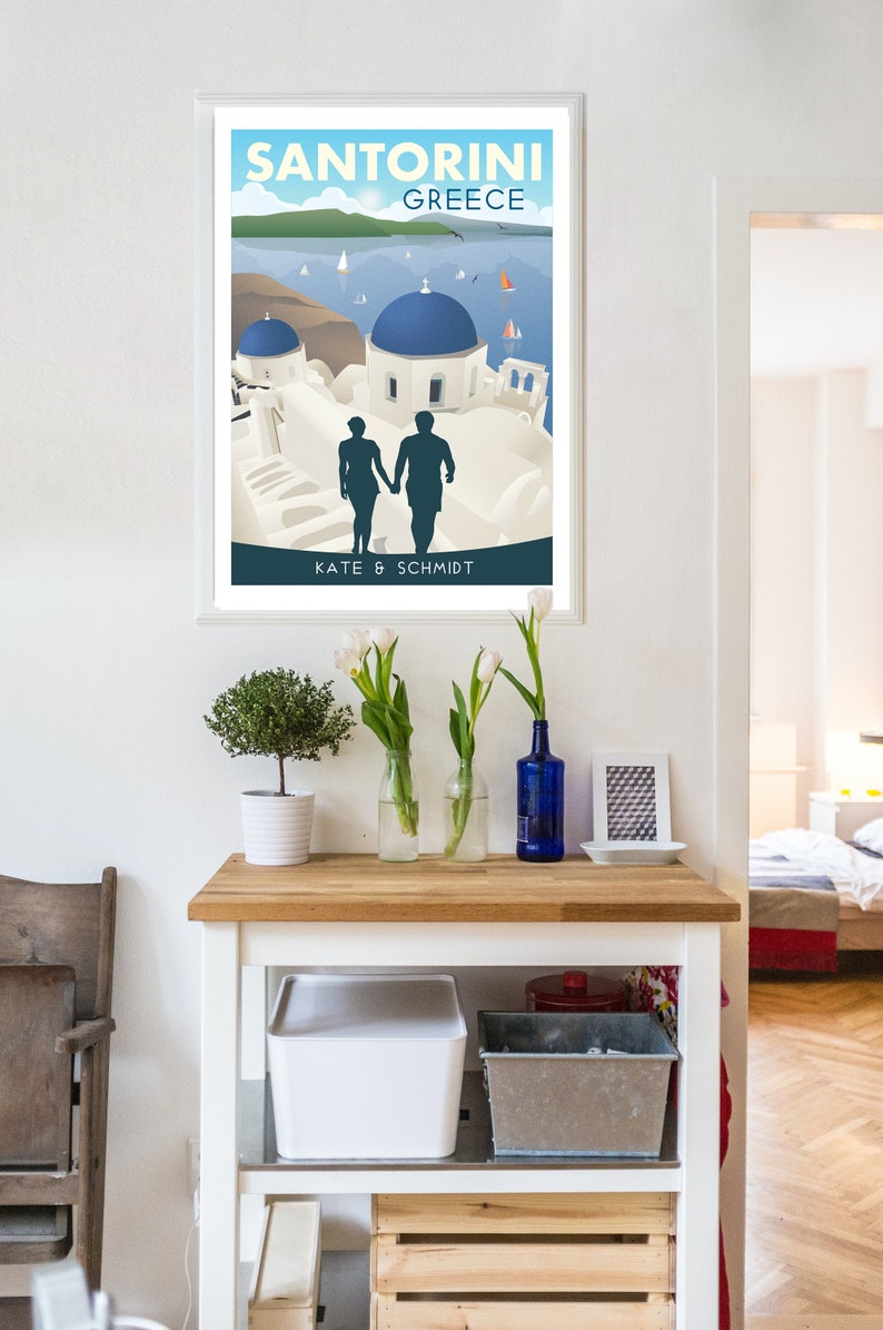 May include: A white framed poster with a colorful illustration of Santorini, Greece. The poster features a couple walking hand-in-hand in front of white buildings with blue domes. The text "Santorini Greece" is at the top of the poster and "Kate & Schmidt" is at the bottom.