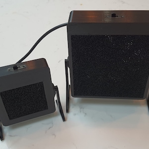 Könnte beinhalten: Zwei schwarze, rechteckige Lautsprecher mit einer strukturierten, dunkelgrauen Oberfläche. Jeder Lautsprecher hat einen schwarzen Ständer und einen Schalter oben. Ein schwarzes Kabel verbindet die Lautsprecher. Die Lautsprecher sind unterschiedlich groß.