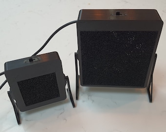May include: Two black, rectangular speakers with a textured, dark gray surface. Each speaker has a black stand and a switch on top. A black cable connects the speakers. The speakers are of different sizes.