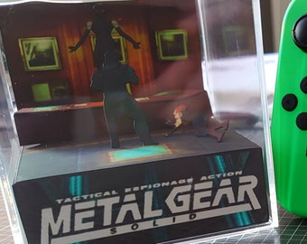 May include: A green Nintendo Switch console with a black screen displaying the Animal Crossing: New Horizons game icon. The console is next to a clear acrylic box with a 3D diorama of the Metal Gear Solid video game. The text "Tactical Espionage Action Metal Gear Solid" is printed on the base of the diorama. The text "The Perfect Gift!" is printed below the diorama.