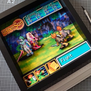 Può includere: Un diorama 3D incorniciato di una scena di battaglia di videogioco di Golden Sun. La scena presenta i personaggi Isaac e Garet in un ambiente boschivo. I personaggi sono pixelati e hanno un effetto 3D. Il diorama è incorniciato in nero e ha una parte anteriore in vetro. Il testo "Golden Sun" è visibile nell'angolo in alto a sinistra del diorama. Il testo "Fight" è visibile nell'angolo in basso a destra del diorama.
