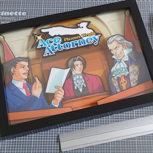 Könnte beinhalten: Gerahmtes Kunstwerk mit Charakteren aus "Ace Attorney". Die Szene zeigt einen Gerichtssaal mit drei Figuren, der Titel "Ace Attorney" ist oben zu sehen. Ein Lineal, zwei Teppichmesser und ein Papageienaufkleber sind ebenfalls sichtbar.