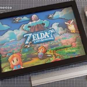 Könnte beinhalten: Gerahmtes Kunstwerk mit einem geschichteten Papier-Diorama des Videospiels "The Legend of Zelda: Link's Awakening". Die Szene zeigt Charaktere, einen Vulkan und das Logo des Spiels. Ein Lineal, ein Papageienaufkleber und Bastelwerkzeuge liegen daneben.