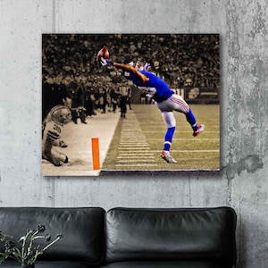 May include: A canvas print of an American football game scene. A player in a blue uniform leaps to catch a football, with a sepia-toned crowd and field in the background. The image is mounted on a gray wall.