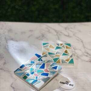 May include: Two square mosaic coasters with a geometric pattern of blue, green, pink, and gold tiles. The coasters are on a white marble surface with a sticker that says "The Crafty Kit".