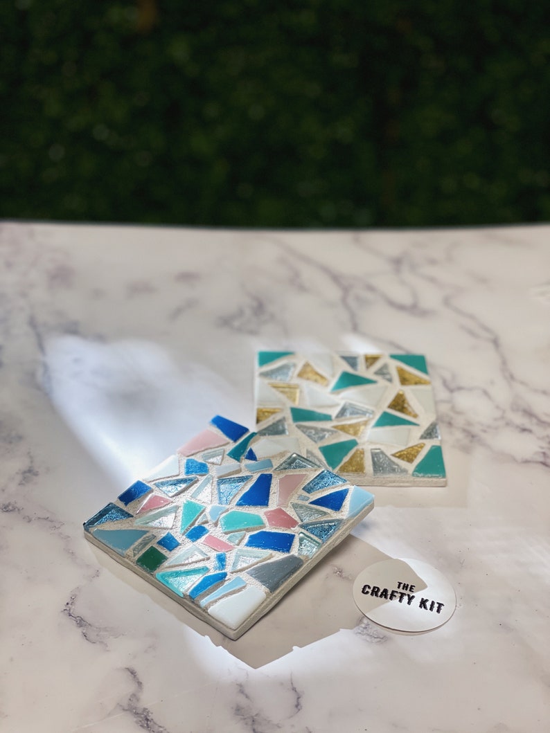 May include: Two square mosaic coasters with a geometric pattern of blue, green, pink, and gold tiles. The coasters are on a white marble surface with a sticker that says "The Crafty Kit".
