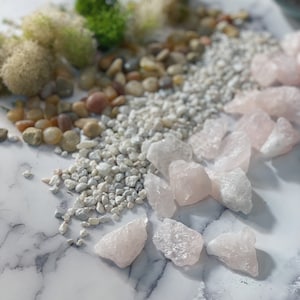 Pode incluir: Uma cole&ccedil;&atilde;o de pequenos cristais de quartzo rosa claro sobre uma superf&iacute;cie de m&aacute;rmore branco. Os cristais est&atilde;o espalhados ao redor de um monte de cascalho branco.