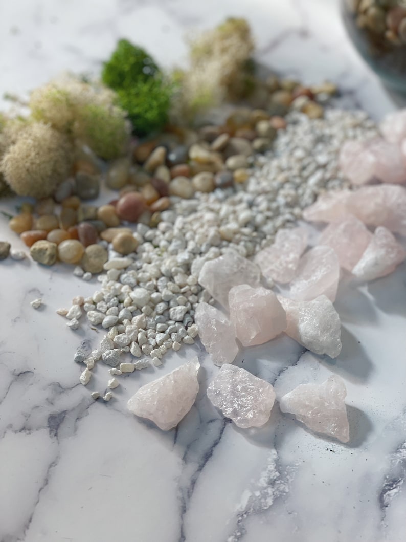 Pode incluir: Uma cole&ccedil;&atilde;o de pequenos cristais de quartzo rosa claro sobre uma superf&iacute;cie de m&aacute;rmore branco. Os cristais est&atilde;o espalhados ao redor de um monte de cascalho branco.