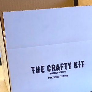 May include: A white cardboard box with the text "THE CRAFTY KIT" in black lettering. The text "TOGETHER WE GROW" is printed below the logo, followed by the website address "WWW.THECRAFTYKIT.COM".