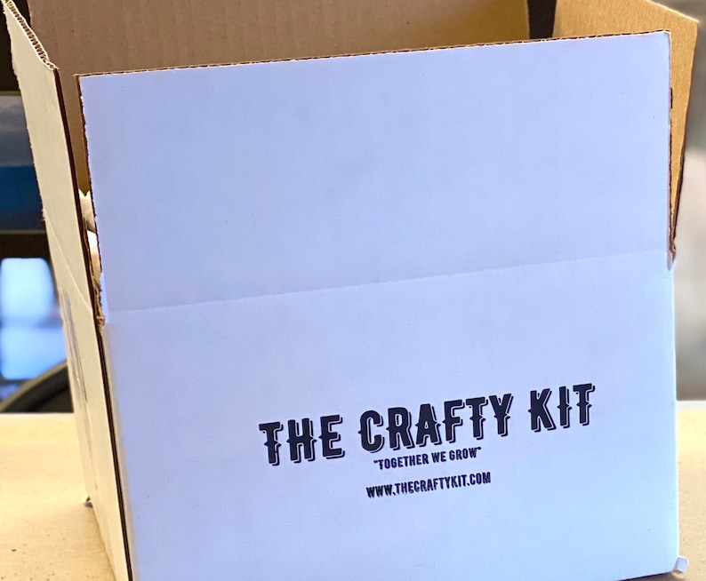 May include: A white cardboard box with the text "THE CRAFTY KIT" in black lettering. The text "TOGETHER WE GROW" is printed below the logo, followed by the website address "WWW.THECRAFTYKIT.COM".