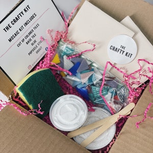 May include: A cardboard box filled with supplies for a mosaic craft kit. The kit includes colored glass tiles, glue, grout, a sponge, and a base tile. The box is decorated with pink tissue paper and a white sticker that says "The Crafty Kit".
