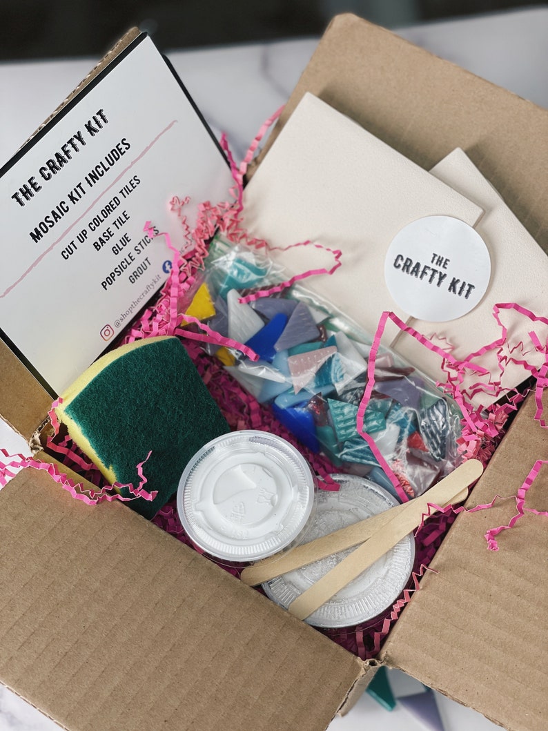 May include: A cardboard box filled with supplies for a mosaic craft kit. The kit includes colored glass tiles, glue, grout, a sponge, and a base tile. The box is decorated with pink tissue paper and a white sticker that says "The Crafty Kit".