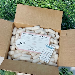 Pode incluir: Uma caixa de cart&atilde;o castanho cheia de amendoins de embalagem brancos. Um cart&atilde;o rosa e branco com o texto "Voc&ecirc; foi convidado para um evento especial!" e um autocolante com o texto "The Crafty Kit" s&atilde;o vis&iacute;veis.