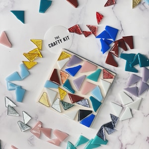 May include: A close-up of a mosaic tile kit with a variety of colors and shapes. The kit includes a small square tile with a mosaic design, as well as a variety of loose tiles in different colors and shapes. The kit is labeled "The Crafty Kit".