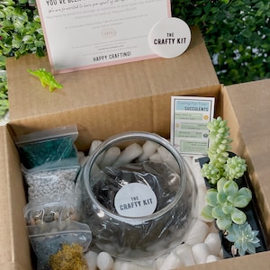 Pode incluir: Uma caixa de cart&atilde;o castanho cheia de suprimentos para um kit de terr&aacute;rio de suculentas. A caixa cont&eacute;m um globo de vidro transparente, terra, pedras, musgo e uma pequena planta suculenta. A caixa tamb&eacute;m inclui um cart&atilde;o com o texto "The Crafty Kit" e instru&ccedil;&otilde;es para cuidar da suculenta.