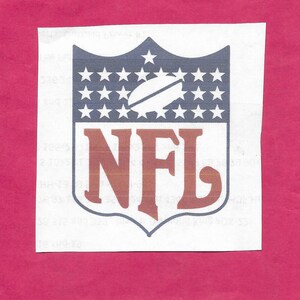 May include: The NFL logo, a shield-shaped emblem, features a football and stars on a blue background, with the letters "NFL" in brown. The logo is printed on a white, rectangular piece of paper, set against a pink background.