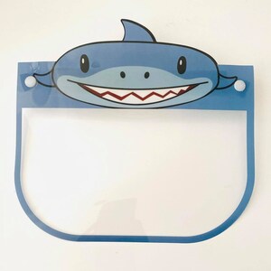 May include: A blue plastic face shield with a cartoon shark design. The shield has a clear plastic visor and two white buttons on the sides.