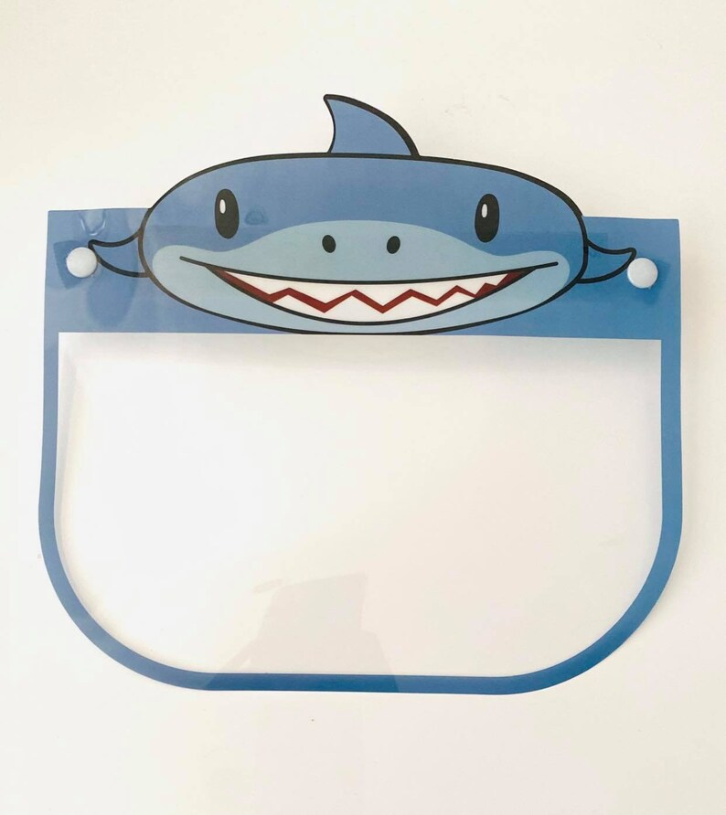 May include: A blue plastic face shield with a cartoon shark design. The shield has a clear plastic visor and two white buttons on the sides.