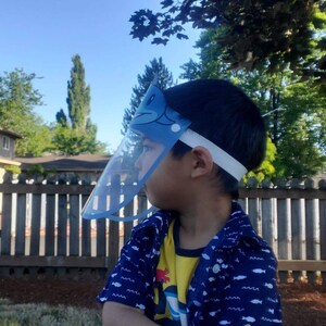 May include: A young child wearing a blue and white face shield with a cartoon fish design. The child is wearing a blue and yellow shirt with a white fish pattern. The child is standing in a grassy area with a wooden fence in the background.