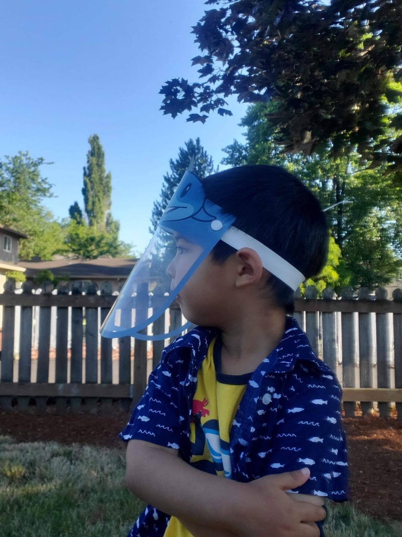 May include: A young child wearing a blue and white face shield with a cartoon fish design. The child is wearing a blue and yellow shirt with a white fish pattern. The child is standing in a grassy area with a wooden fence in the background.