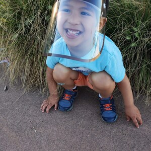 May include: A young child wearing a blue shirt and a clear plastic face shield with a black bear design. The child is smiling and crouching down on a brown surface.