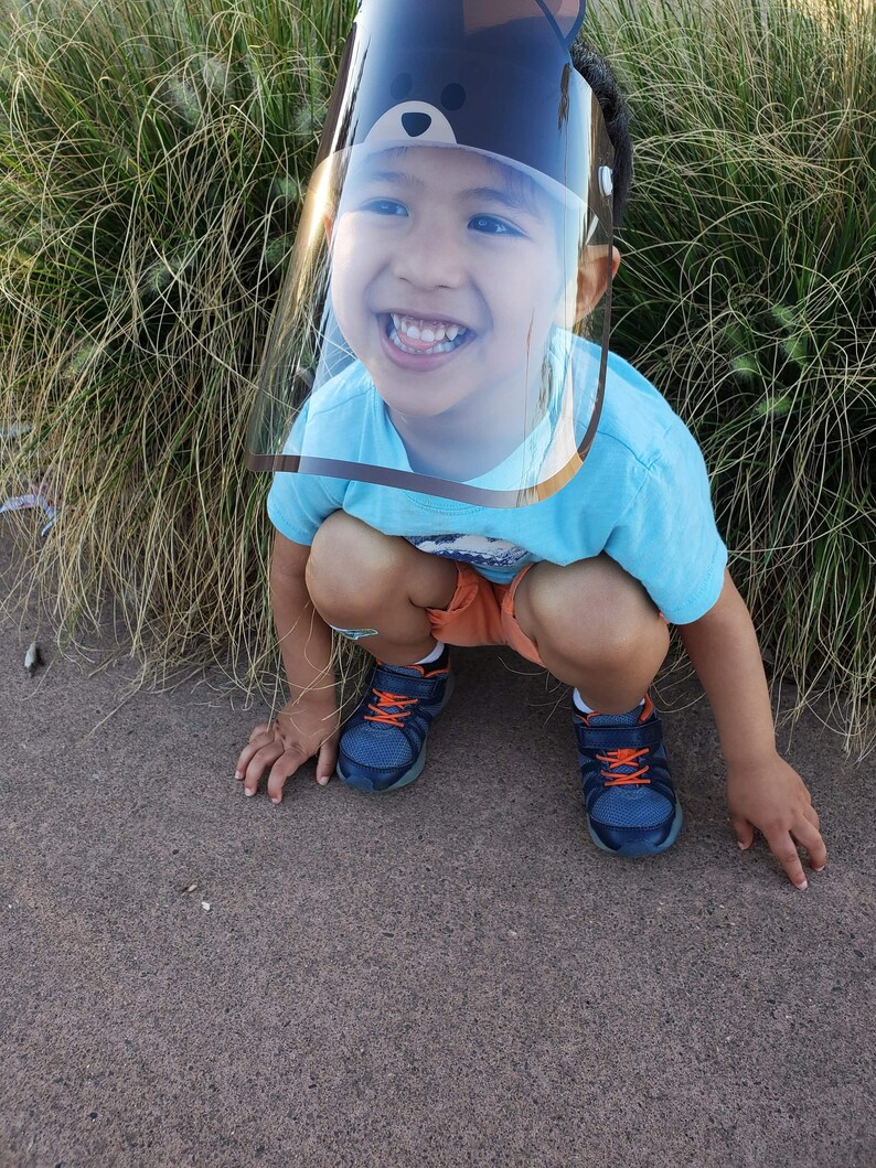 May include: A young child wearing a blue shirt and a clear plastic face shield with a black bear design. The child is smiling and crouching down on a brown surface.