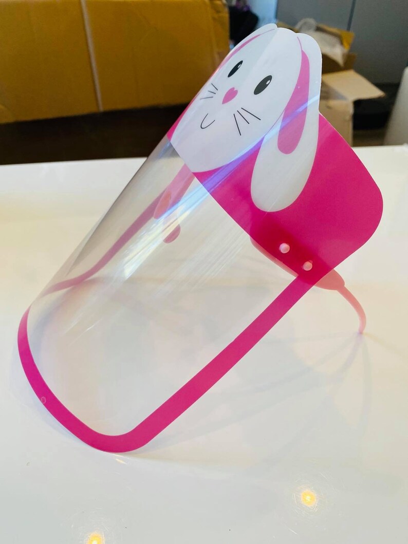 May include: Pink plastic face shield with a clear visor and a white bunny design. The bunny has black eyes and a pink nose.