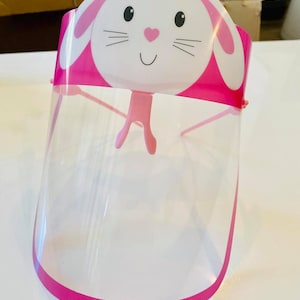 May include: Pink plastic face shield with a white bunny design. The bunny has black eyes, a pink nose, and a pink mouth. The shield is clear and has a pink border.