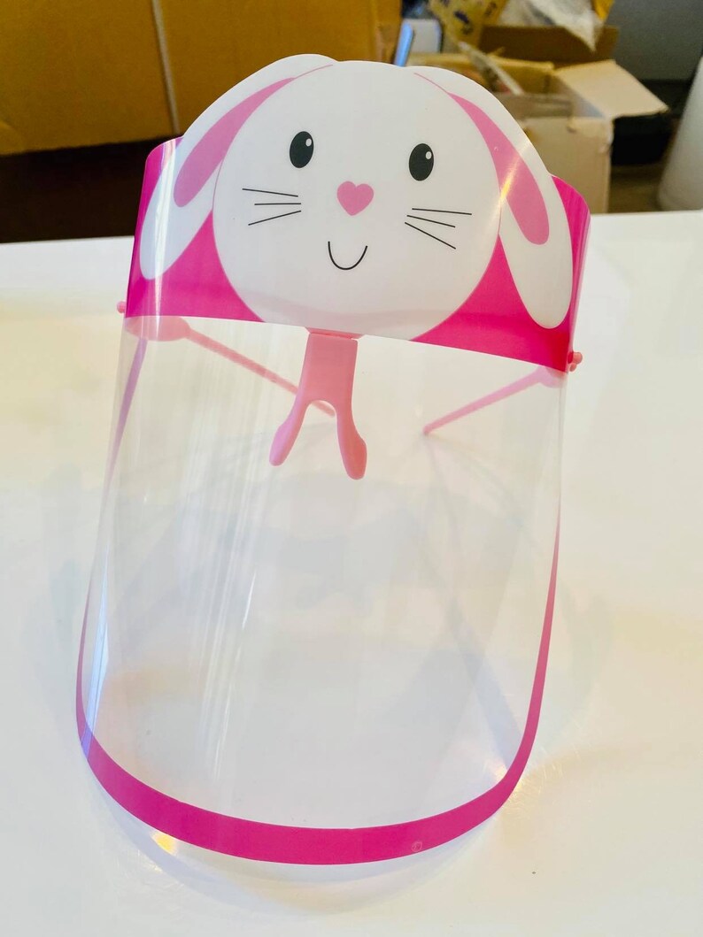 May include: Pink plastic face shield with a white bunny design. The bunny has black eyes, a pink nose, and a pink mouth. The shield is clear and has a pink border.