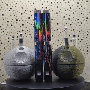 May include: Two silver and black vape pens with colorful Star Wars character designs, sitting on two Death Star-shaped bases. The bases are silver and green.