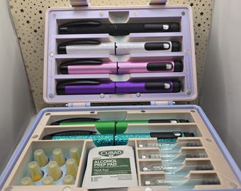 May include: A light purple, two-tiered case with a variety of medical supplies. The top tier holds four colorful injection pens: black, silver, pink, and purple. The bottom tier contains two green and blue pens, alcohol prep pads, and other medical supplies.