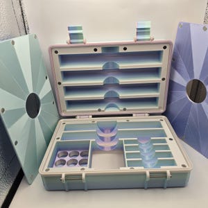 Pu&ograve; includere: Valigetta portaoggetti aperta con pi&ugrave; scomparti in tonalit&agrave; pastello. La valigetta &egrave; azzurra con un bordo rosa e un interno bianco, che rivela ripiani e scomparti. Due pannelli geometrici coordinati sono su entrambi i lati.
