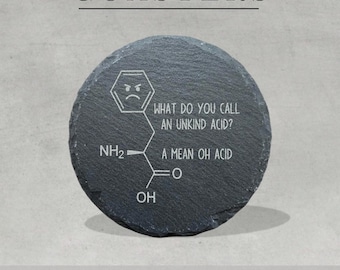 May include: A round, dark gray slate coaster featuring a white engraved chemical structure of an amino acid with an angry face inside the hexagon. The text reads: WHAT DO YOU CALL AN UNKIND ACID? A MEAN OH ACID. The coaster has a rough, natural edge.