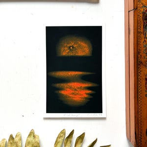 Könnte beinhalten: Ein kleiner Kunstdruck mit dem Titel "Blood Orange" zeigt einen Sonnenuntergang über dem Wasser in Orange- und Schwarztönen. Ein gelber Bleistift und eine Holzkiste sind ebenfalls im Bild, zusammen mit einem Zweig getrockneter Blätter.