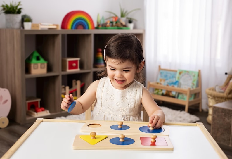 May include: A child is playing with a wooden shape sorter toy. The toy has three sections: a circle, a square, and a triangle. Each section has a corresponding shape that can be inserted into the hole. The child is holding a blue circle shape and is about to insert it into the circle hole. The toy is made of natural wood and has a light brown finish.