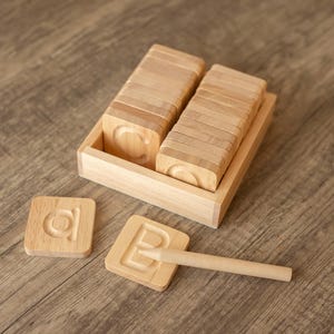 May include: A wooden box filled with letter tiles, with two tiles showing the letters "a" and "b". A wooden stylus is also included.