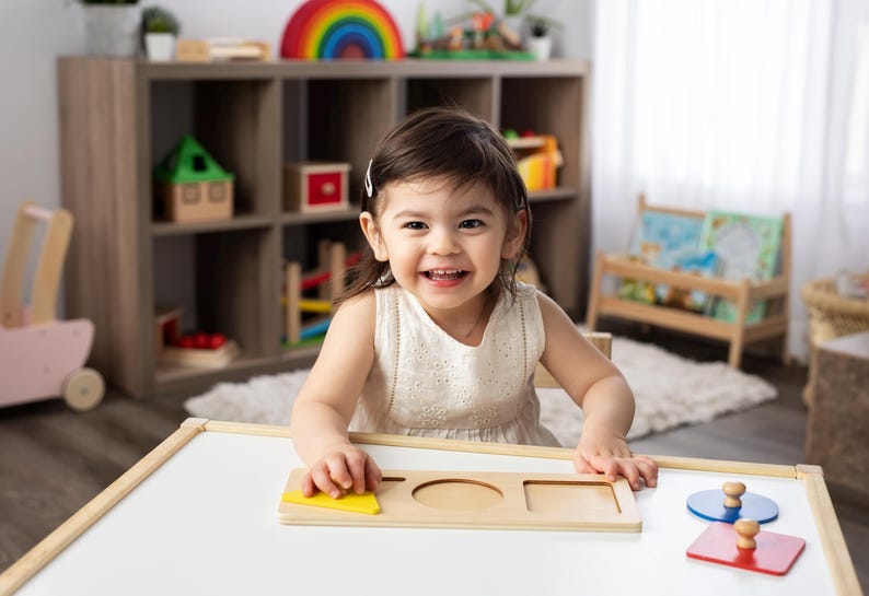 May include: A young child sits at a table playing with a wooden shape puzzle. The puzzle has a yellow triangle, a circle, a square, and a rectangle. The child is smiling and appears to be enjoying the activity.