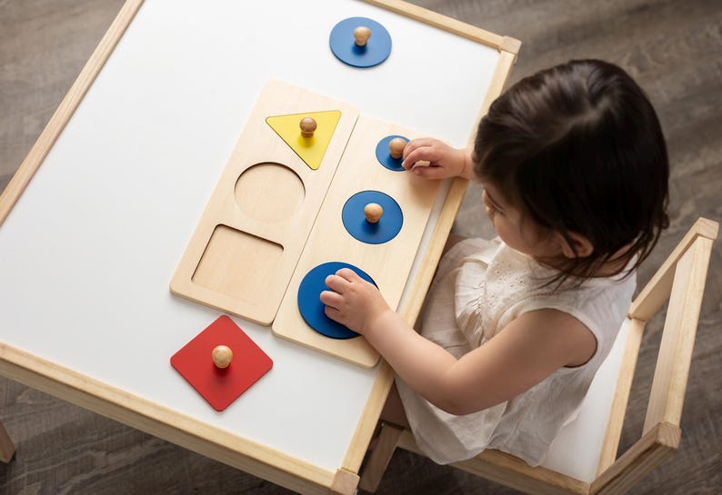 May include: A child is playing with a wooden shape sorter toy. The toy has different coloured shapes, including a yellow triangle, a red square, and blue circles. The child is matching the shapes to the corresponding holes in the board.