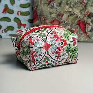 Pode incluir: Uma pequena bolsa de cosméticos quadrada com zíper vermelho e estampa floral. A bolsa apresenta um fundo branco com bagas vermelhas, folhas verdes e detalhes florais vermelhos. A bolsa tem aproximadamente 10 cm de largura e 10 cm de altura.