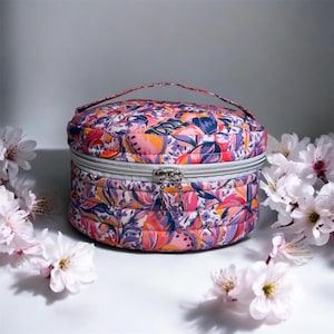 Op de afbeelding: Een ronde, gewatteerde make-up tas met een kleurrijk bloemenpatroon in roze, paars, oranje en blauw. De tas heeft een zilveren rits, een klein handvat en is omgeven door witte bloemen.