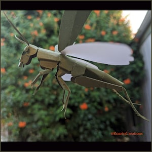 May include: A papercraft grasshopper with green wings and a white underbelly. The grasshopper is posed in flight with its legs extended. The background is a blurry image of green foliage with orange flowers.