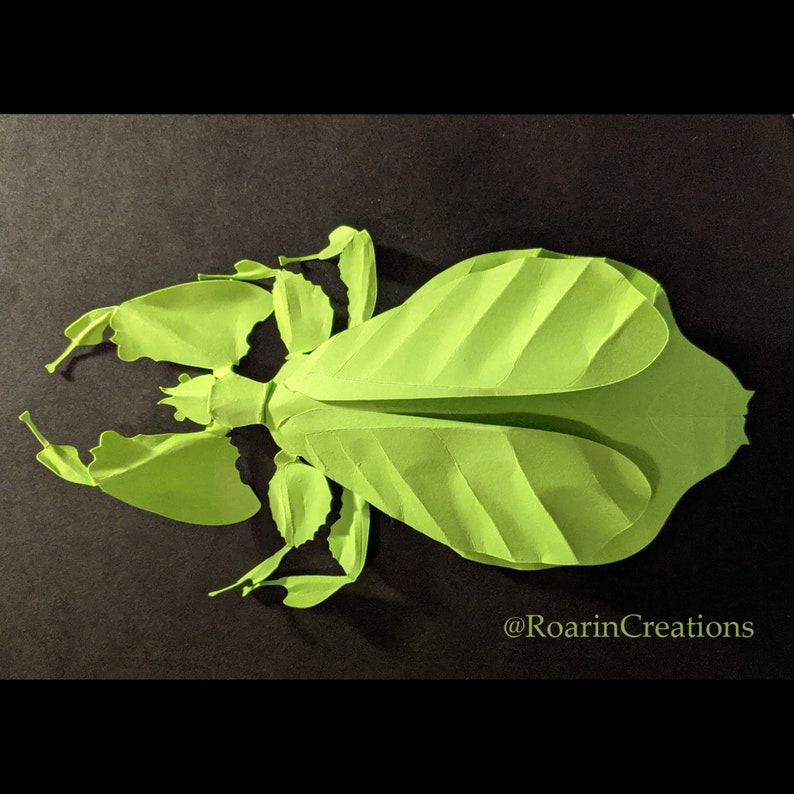 Pu&ograve; includere: Un modello di insetto stecco in carta verde, con dettagli intricati e un aspetto realistico. Il modello &egrave; in posa con le gambe distese, che ricordano un vero insetto. Il nome utente Instagram dell'artista, @RoarincCreations, &egrave; visibile nell'angolo in basso a destra.