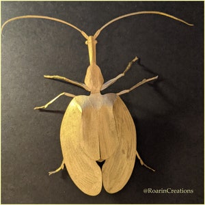 May include: A gold-colored paper insect with long antennae and a detailed body. The insect is on a black background. @RoarInCreations