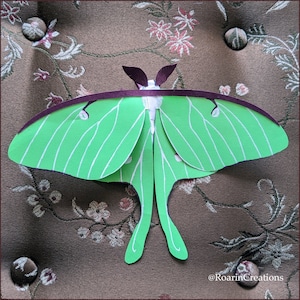 May include: A green and white paper cutout of a Luna moth with purple accents. The moth is on a brown fabric background with floral patterns.
