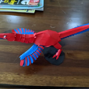 May include: A handcrafted paper bird model with a red body and blue and red wings, set on a black circular base. The papercraft bird is made from folded paper.