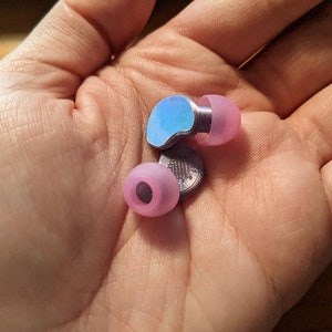 以下が含まれることがあります： メタリックシルバーのステムと虹色のブルーのトップを備えたイヤホン。イヤーチップはソフトピンク色です。イヤホンは小さく、耳に快適にフィットするように設計されています。